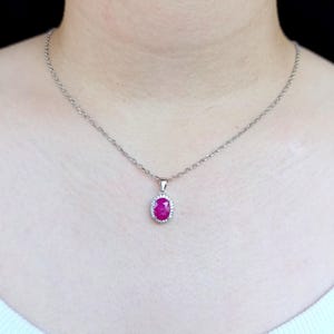 May include: A silver-toned chain necklace featuring an oval-shaped, vibrant pinkish-red gemstone pendant. The central stone is surrounded by a halo of small, clear, sparkling stones set in a metallic frame, suspended from a delicate link chain.