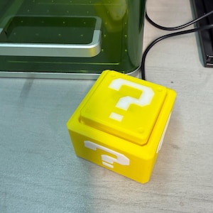 Può includere: Un cubo giallo brillante con un punto interrogativo bianco sulla parte superiore e laterale. Il cubo è appoggiato su una superficie grigio chiaro. Il cubo è una replica del blocco punto interrogativo della serie di videogiochi Super Mario.