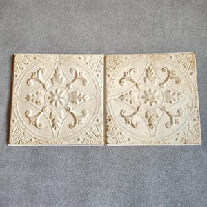 May include: Two square off-white metal ceiling tiles featuring a raised embossed floral pattern with a central rosette, curved scrollwork, and geometric corner accents. The surface has a distressed, weathered finish with visible brown patina.