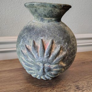 May include: A round, dark green ceramic vase with a wide, flared rim. The exterior features a prominent, raised relief design of a sun face with radiating rays. The surface has a weathered, matte texture with subtle hints of reddish-brown undertones.