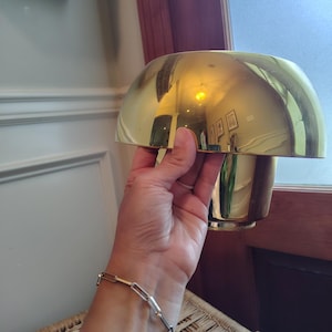 May include: A polished gold-tone metal lamp with a wide, dome-shaped shade and a cylindrical base. The reflective surface of the lamp shows a clear mirror-like finish, capturing the surrounding environment in its curved, smooth metallic exterior.