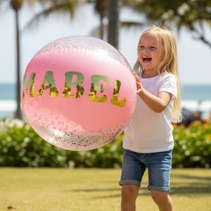 May include: A clear, inflatable beach ball featuring a pink center panel with the name MABEL printed in large, metallic gold capital letters. The transparent sections of the ball are filled with small, scattered gold glitter particles.