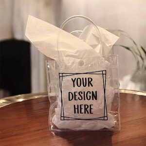 May include: Clear plastic gift bag with a handle, filled with white tissue paper. The bag has a black square frame with the words "YOUR DESIGN HERE" printed in black. The bag is sitting on a wooden surface.