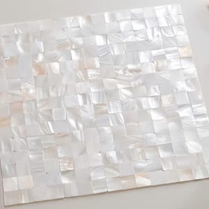 Puede incluir: Panel decorativo cuadrado, compuesto por numerosas baldosas blancas iridiscentes dispuestas en mosaico. Las baldosas tienen un brillo nacarado sutil, creando una superficie texturizada. El panel se encuentra sobre un fondo liso y claro. Se ve una espiga de trigo en la esquina superior derecha.