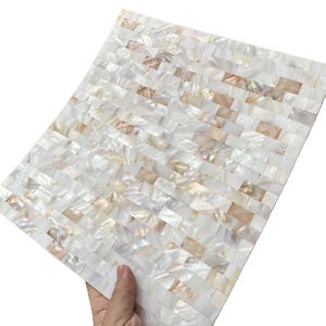 Peut inclure: Une feuille de carrelage en mosaïque rectangulaire composée de petits morceaux rectangulaires de nacre irisée. Les carreaux sont disposés en motif de briques, avec un mélange de teintes blanches et marron clair. La feuille est tenue par une main.