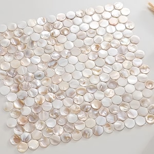 May include: A square mosaic tile sheet composed of numerous small, round, iridescent mother-of-pearl tiles. The tiles vary in color, with shades of white, cream, and subtle hints of gold. The overall effect is a shimmering, textured surface.