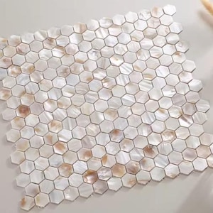 Puede incluir: Un cuadrado de baldosas de mosaico hexagonales. Las baldosas son principalmente blancas con acentos iridiscentes y bronceados. El mosaico está dispuesto en un patrón de panal, creando una superficie visualmente atractiva. Las baldosas probablemente están hechas de materiales naturales.