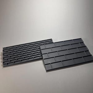 May include: Two gray plastic molds with a brick pattern. One mold has a more detailed brick layout, while the other has a simpler design. The molds are likely used for creating decorative elements or textures.