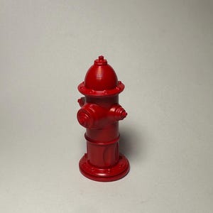 Può includere: Una miniatura di un idrante rosso brillante. L'idrante ha una parte superiore arrotondata, due ugelli laterali e una base circolare. Il design è una rappresentazione classica di un idrante, comune negli ambienti urbani. L'idrante è di un unico colore.
