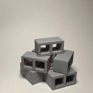 Puede incluir: Una pequeña pila de bloques de hormigón grises con dos agujeros rectangulares. Los bloques están apilados, con un poco de pintura roja visible en el interior de algunos. El fondo es de color blanco roto.