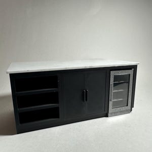 Puede incluir: Una isla de cocina con encimera de mármol blanco y base negra mate. La unidad cuenta con tres estantes abiertos a la izquierda, un armario central de doble puerta con tiradores verticales y un enfriador de bebidas con puerta de cristal a la derecha.