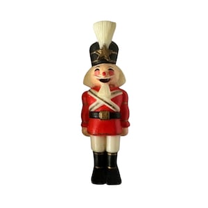 May include: A nutcracker figurine wearing a black hat with a gold star, a red jacket with white cross-straps, and black boots. The figure has a white beard, white hair, and a painted face with rosy cheeks, a red nose, and an open mouth.