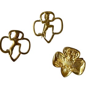 May include: Three vintage gold-toned Girl Scout pins. Two pins feature a figure within a trefoil shape, and the third pin displays an eagle emblem. These pins are likely collectible items, showcasing classic scouting imagery.