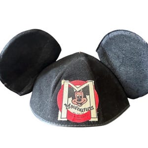 May include: Black Mickey Mouse ears hat with a red and white Mouseketeers logo. The logo features Mickey Mouse and the text "Mouseketeers" and "1955 Disney Club". The hat has two large, rounded ears.