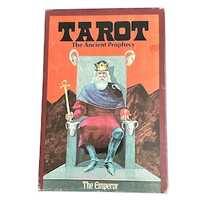 May include: A vintage Tarot card deck box with the title "TAROT" in bold black letters. The image depicts "The Emperor" seated on a throne, holding a staff. The background is a reddish-orange with mountains.