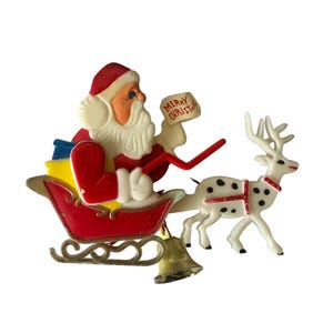 May include: A vintage Christmas brooch featuring Santa Claus in a red sleigh pulled by a white reindeer with black spots. Santa holds a sign that reads "Merry Christmas". The sleigh has a gold bell.