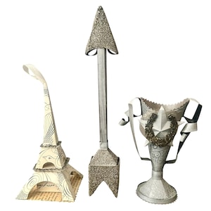 May include: Three decorative paper sculptures: an Eiffel Tower, an arrow, and a trophy. The Eiffel Tower is white with black details and glitter accents. The arrow has a glittered arrowhead and base. The trophy is silver with a star and wreath.