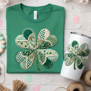 May include: A green t-shirt and a white tumbler, both with a four-leaf clover design. The clover is made from various green fabrics with patterns like gingham and floral. The tumbler has a silver lid. The items are on a white surface.