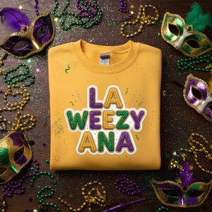 Può includere: Felpa giallo senape con la scritta "LA WEEZY ANA" in un design colorato e strutturato. L'immagine è circondata da maschere, perline e coriandoli del Mardi Gras, creando un'atmosfera festosa.
