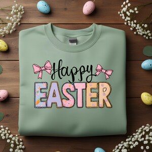 May include: A sage green sweatshirt featuring the text "Happy Easter". The word "Easter" is in colourful, patterned letters, with pink bows above and below the text. The image is decorated with Easter eggs and flowers.