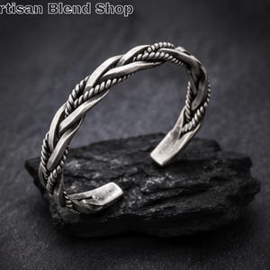 May include: A silver cuff bracelet with a braided rope design. The open cuff is displayed on a dark, textured surface. The text "Artisan Blend Shop" is visible in the upper left corner.