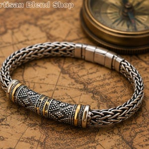 May include: Silver-toned bracelet with a woven chain design and a decorative center section. The center features a detailed pattern with gold accents. The bracelet is displayed on a map.
