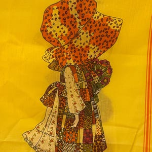 May include: A yellow fabric with a repeating pattern of a girl in a patchwork dress and bonnet. The girl is holding flowers. The design includes orange and brown accents, with the words "HOLLY HOBBIE" at the bottom of the image.