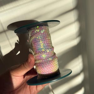 May include: A spool of iridescent sequins, wrapped around a dark teal disc. The sequins shimmer with pink, yellow, and green hues. The spool is held in a hand, with the light creating shadows on the wall.