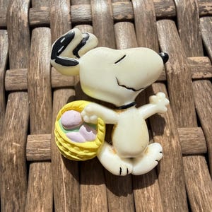 May include: A Snoopy figurine in a running pose, holding a yellow basket with pastel-colored Easter eggs. The figurine is white with black accents on the ears and feet. The background is a woven brown surface.