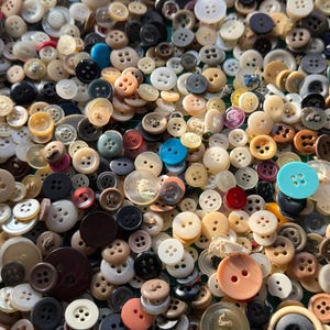May include: A collection of various buttons in different sizes, shapes, and colors. The buttons range from small to large, with colors including white, black, brown, beige, and a few pops of blue and red. Some buttons have two holes, while others have four.
