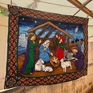 May include: A rectangular woven tapestry featuring a nativity scene with a central stable, figures of Mary, Joseph, and the infant Jesus, accompanied by an angel, sheep, and a donkey, all framed by a decorative border with red and gold floral motifs.