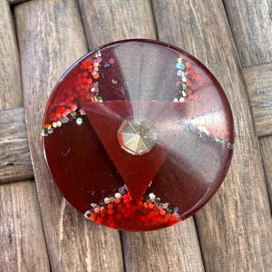 May include: A round, dark red resin object with a triangular design and a central, faceted gem. The resin is embedded with red and iridescent glitter, creating a sparkling effect. The object is set against a textured, wooden surface.