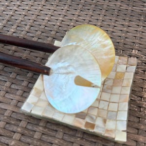 May include: A pair of dark brown wooden hair sticks featuring circular, iridescent mother-of-pearl accents with a small triangular cutout. The hair sticks are resting on a square tray covered in a mosaic of small, light-colored shell tiles.