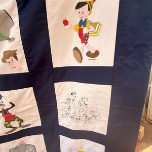 Peut inclure: Une courtepointe bleu marine avec des personnages de dessins anim&eacute;s peints &agrave; la main sur des carr&eacute;s blancs. Pinocchio tient une pomme, tandis que d'autres carr&eacute;s repr&eacute;sentent Dumbo, des chiens dalmatiens et Woody. La courtepointe est un patchwork avec des illustrations color&eacute;es.