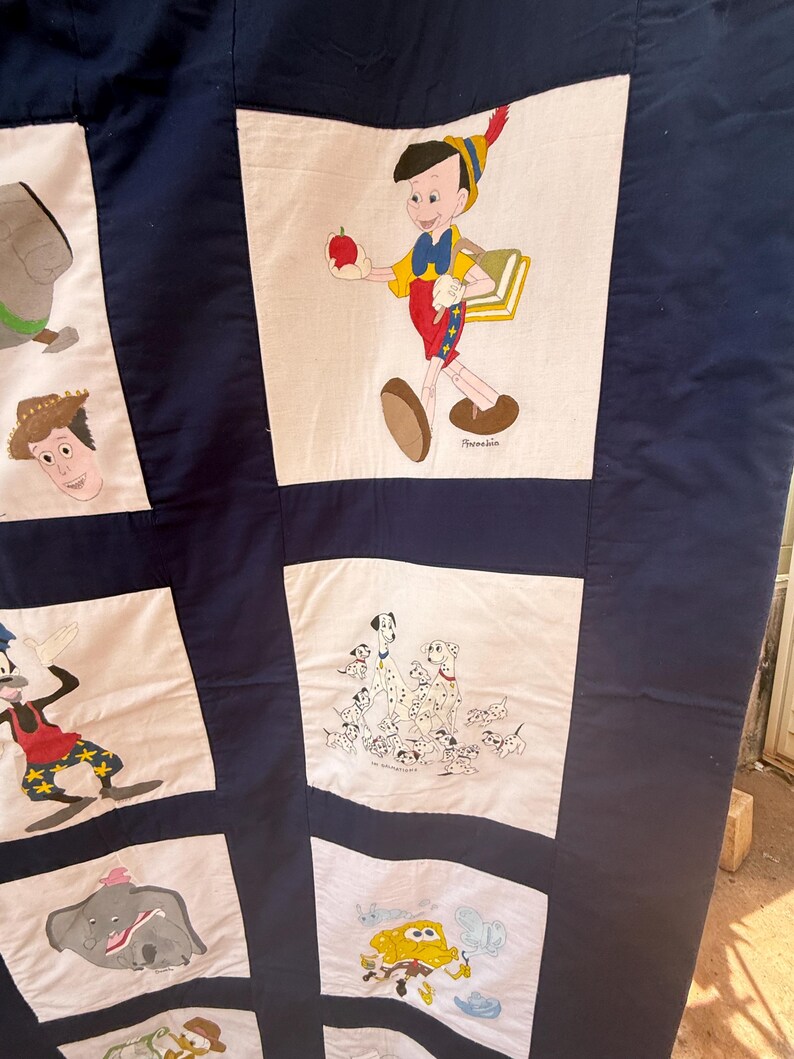 Peut inclure: Une courtepointe bleu marine avec des personnages de dessins anim&eacute;s peints &agrave; la main sur des carr&eacute;s blancs. Pinocchio tient une pomme, tandis que d'autres carr&eacute;s repr&eacute;sentent Dumbo, des chiens dalmatiens et Woody. La courtepointe est un patchwork avec des illustrations color&eacute;es.