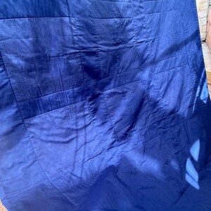 Peut inclure: Gros plan sur une couverture matelass&eacute;e bleu marine. La couverture est faite d'un tissu lisse et brillant et pr&eacute;sente un motif cousu de formes rectangulaires. La couverture est pli&eacute;e et la lumi&egrave;re du soleil cr&eacute;e des ombres sur la surface.