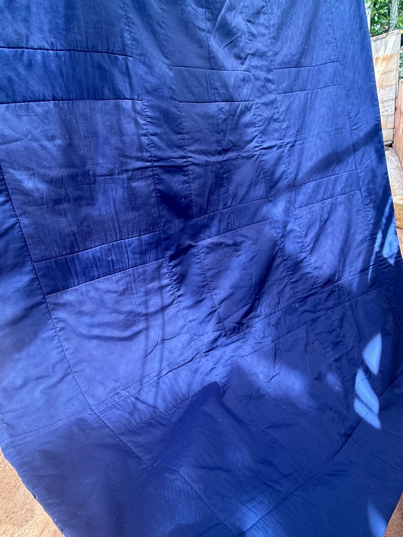 Peut inclure: Gros plan sur une couverture matelass&eacute;e bleu marine. La couverture est faite d'un tissu lisse et brillant et pr&eacute;sente un motif cousu de formes rectangulaires. La couverture est pli&eacute;e et la lumi&egrave;re du soleil cr&eacute;e des ombres sur la surface.
