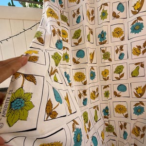 May include: A white fabric featuring a repeating grid pattern of various botanical illustrations including yellow, blue, and green flowers and green pears. The text AMERITEX CO. is printed in black along the edge of one of the floral squares.