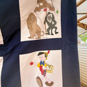 Peut inclure: Une courtepointe faite &agrave; la main avec des illustrations de dessins anim&eacute;s sur des panneaux. Un panneau montre Mufasa, un lion, et d'autres personnages du Roi Lion. Un autre panneau repr&eacute;sente Pinocchio tenant une pomme. La courtepointe a une bordure bleu marine et des carr&eacute;s blancs.