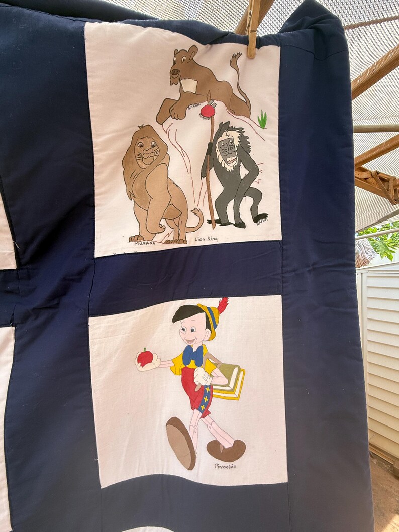 Peut inclure: Une courtepointe faite &agrave; la main avec des illustrations de dessins anim&eacute;s sur des panneaux. Un panneau montre Mufasa, un lion, et d'autres personnages du Roi Lion. Un autre panneau repr&eacute;sente Pinocchio tenant une pomme. La courtepointe a une bordure bleu marine et des carr&eacute;s blancs.