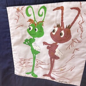 Peut inclure: Un tissu bleu marine avec un patch carr&eacute; blanc repr&eacute;sentant une illustration de dessin anim&eacute; de deux personnages ressemblant &agrave; des insectes. L'un est vert, l'autre est marron. Les mots "Bugs Life" sont &eacute;crits en bas &agrave; droite.