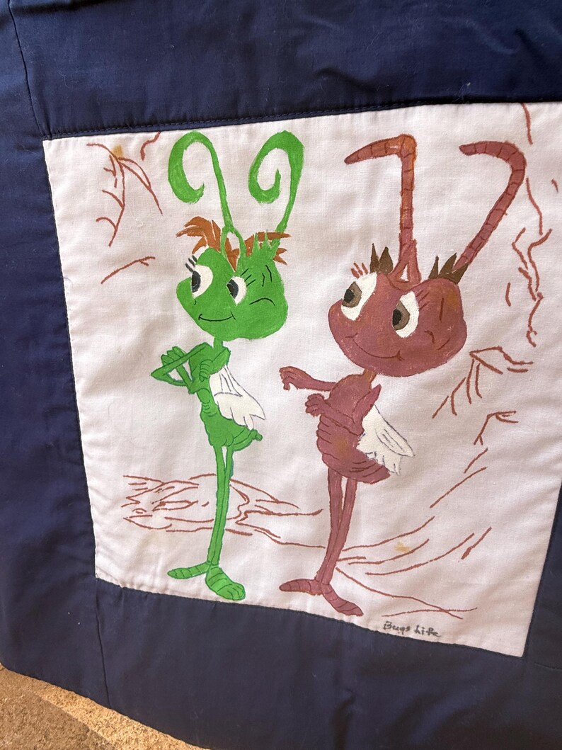 Peut inclure: Un tissu bleu marine avec un patch carr&eacute; blanc repr&eacute;sentant une illustration de dessin anim&eacute; de deux personnages ressemblant &agrave; des insectes. L'un est vert, l'autre est marron. Les mots "Bugs Life" sont &eacute;crits en bas &agrave; droite.