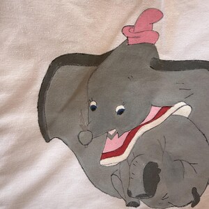 Peut inclure: Illustration peinte &agrave; la main de Dumbo l'&eacute;l&eacute;phant, avec un corps gris, de grandes oreilles et un chapeau rose. L'&oelig;uvre comprend le nom "Dumbo" dans un style manuscrit, sur fond blanc.