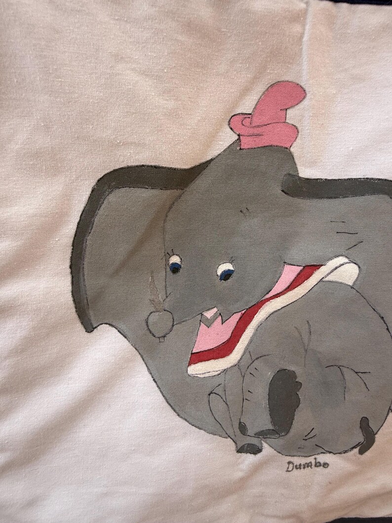 Peut inclure: Illustration peinte &agrave; la main de Dumbo l'&eacute;l&eacute;phant, avec un corps gris, de grandes oreilles et un chapeau rose. L'&oelig;uvre comprend le nom "Dumbo" dans un style manuscrit, sur fond blanc.