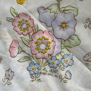 May include: White fabric with a floral pattern featuring pink, blue, and purple flowers, green leaves, and a yellow butterfly. The text "©1983 OAA, INC." is printed on the fabric. This fabric is suitable for sewing projects.