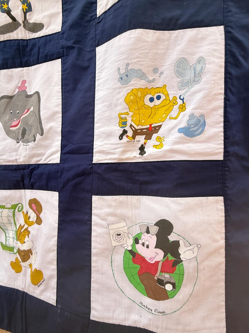 Peut inclure: Une courtepointe faite &agrave; la main avec des personnages de dessins anim&eacute;s tels que Dumbo, Bob l'&eacute;ponge et Mickey Mouse. La courtepointe a un fond bleu marine avec des carr&eacute;s blancs pr&eacute;sentant les personnages. Les personnages sont de couleurs vari&eacute;es, dont le jaune, le gris et le rouge.