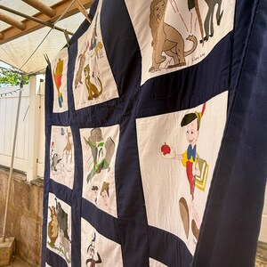 Peut inclure: Une courtepointe faite &agrave; la main avec divers personnages de dessins anim&eacute;s sur des carr&eacute;s blancs, encadr&eacute;s de tissu bleu marine. La courtepointe est suspendue &agrave; l'ext&eacute;rieur, pr&eacute;sentant un motif patchwork avec des images de Pinocchio, Simba et d'autres personnages.
