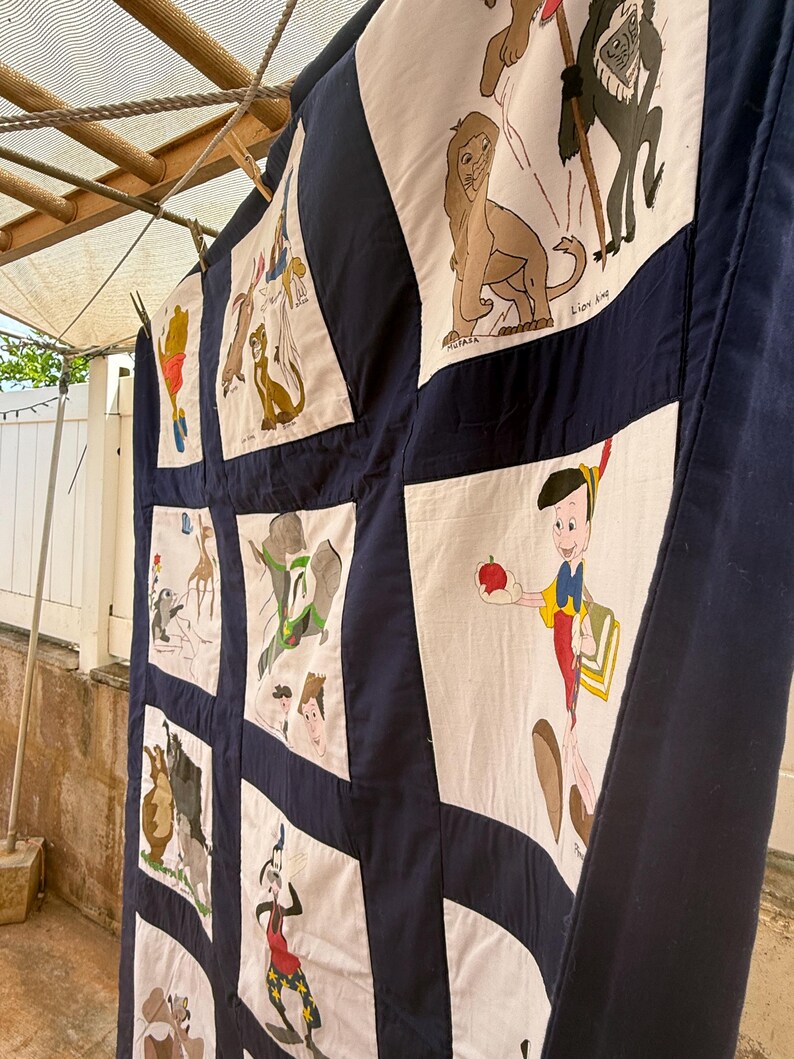 Peut inclure: Une courtepointe faite &agrave; la main avec divers personnages de dessins anim&eacute;s sur des carr&eacute;s blancs, encadr&eacute;s de tissu bleu marine. La courtepointe est suspendue &agrave; l'ext&eacute;rieur, pr&eacute;sentant un motif patchwork avec des images de Pinocchio, Simba et d'autres personnages.