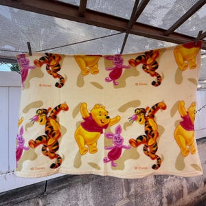 May include: A cream-colored fleece blanket featuring a repeating pattern of Winnie the Pooh, Tigger, and Piglet characters. The design includes the Disney logo printed in small text near the figures, showcasing the iconic yellow, orange, and pink colors.