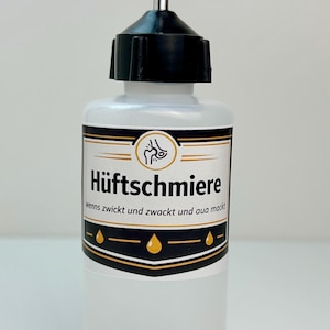Könnte beinhalten: Eine transparente Plastikflasche mit schwarzem Deckel und silberner Düse. Das Etikett zeigt den Text "Hüftschmiere" in fetter Schrift, mit zusätzlichem Text darunter. Das Etikett hat einen goldenen und schwarzen Rand und eine Personengrafik.